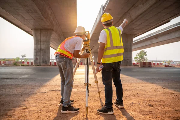 Asian civil engineer and surveyor engineers making measuring and pointing finger under the expressway with theodolite on road works. Survey engineer at road construction site, Surveyor equipment.