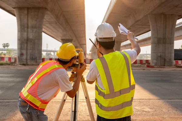 アジア土木技術者と測量士のチームワークは 道路工事のテオドライトで測定する青写真を保持立っています 道路建設現場の測量技術者 測量機器 — ストック写真