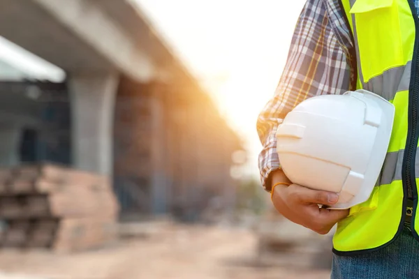 stock image Construction worker or civil engineer holding safety white helmet and construction drawing against the background of road construction site.