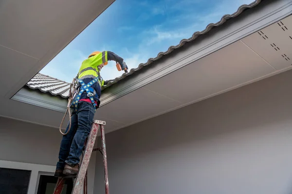 Asian roofer wearing safety harness belt using air or pneumatic nail gun and installing house roof tiles on top new roof at home under construction. Roofing tools.