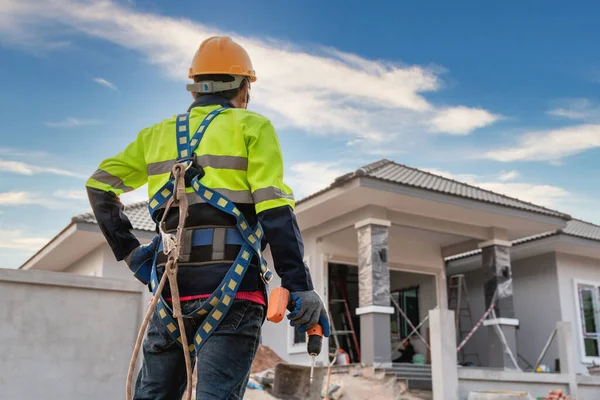 A construction worker stands holding an air gun or pneumatic nail gun and wearing a safety belt for fall protection. the front of the building on the construction site Roofing tools. Repair the roof.