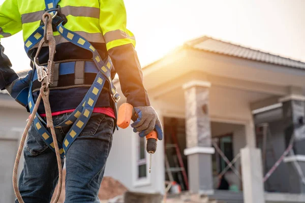 A Roofer wearing safety harness belt stands holding an air gun or pneumatic nail gun. the front of the building on the construction site Roofing tools. Repair the roof.