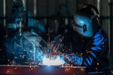 Asyalı işçiler çelik fabrikalarında endüstriyel üniformalar giyip demir maskeler takıyorlar. Fabrikanın içinde kaynak makinesi kullanan endüstriyel işçiler var..
