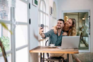 Birlikte selfie çeken çift, dizüstü bilgisayar ve akıllı telefon kullanan mutlu çift. Kafkasyalılar evde, insanlarda ve modern teknoloji konseptinde birlikte eğleniyorlar..