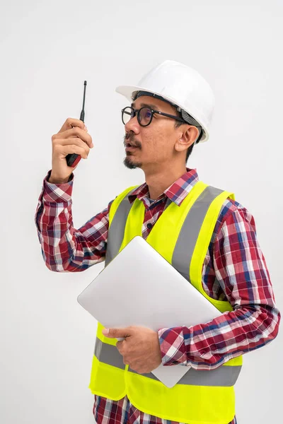 身穿头盔背心的亚洲承包商使用的是带笔记本电脑的对讲机 背景是白色的 建筑承包商概念 垂直图像 — 图库照片