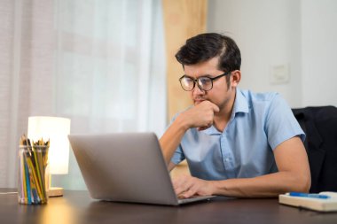 Gözlüklü yakışıklı bir genç masa başında dizüstü bilgisayar kullanıyor sosyal medya bilgilerini okuyor evde kendini geliştirmek için..
