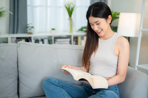 stock image Asian young woman sitting happily reading a book on the sofa in the living room. brain food ideas.