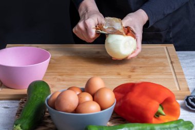 Ahşap mutfak tahtasındaki soğanı soymak için bıçak kullanan bir kadının elleri. Yumurtalı, kırmızı ve yeşil dolmalık biberli, soğanlı ve kabaklı bulanık pembe ve gri kaseler. Yemek hazırlama konsepti. seçici odak