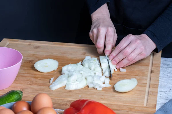 Ahşap mutfak tahtasındaki soğanı kesmek için bıçak kullanan çalışan kadının elleri. Yumurta, kırmızı ve yeşil biber, soğan ve kabaklı bulanık pembe ve gri kaseler. Gıda hazırlama kavramı. Seçmeli odak