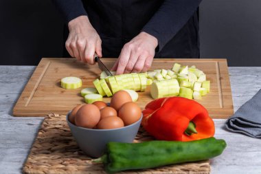Hands of working woman cutting courgette on wooden kitchen board. Faded to front, gray bowl with eggs and red and green pepper. Food preparation concept. selective focu