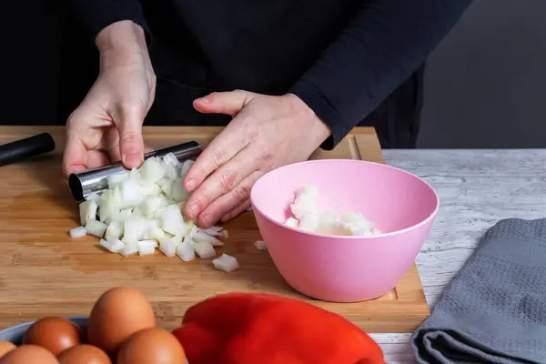 Hands of working woman picking up cut onion on a kitchen board to put it in pink boll. In front of defocused red pepper and gray boll with eggs. Cooking concept. Selective focus