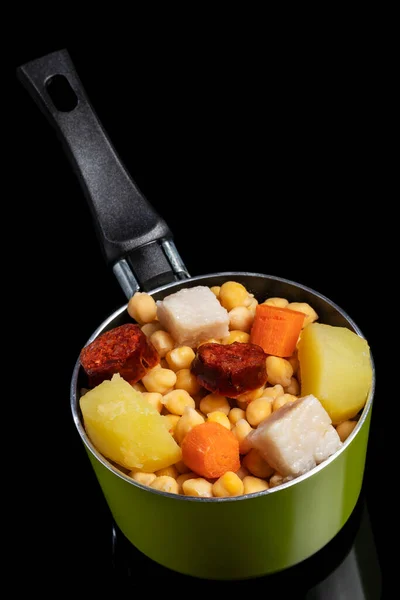 Cooked (Cocido, Guiso) stew, chickpeas with chorizo, bacon, carrot and potato in a green saucepan and on a black background. Concept of traditional Spanish cuisine. Selective focus.