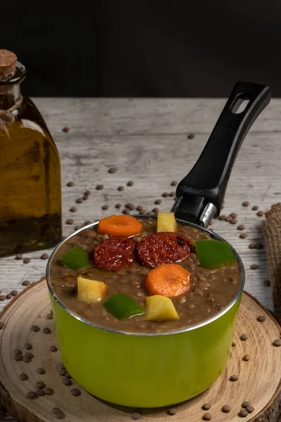 Stewed lentils with chorizo, apple or potato, green pepper and carrot in a green metal saucepan and on a wooden board. Concept of traditional and rustic cooking. selective focus