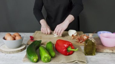Ahşap mutfak tahtasındaki soğanı kesmek için bıçak kullanan çalışan kadının elleri. Yumurtalı gri kase, kırmızı ve yeşil biberli kumaş soğan, kabak ve zeytinyağı şişesi. Gıda hazırlama konsepti.