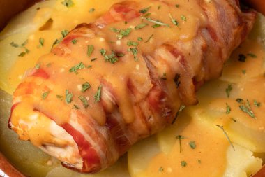 Sosuna domuz pastırması sarılmış peynirle doldurulmuş tavuk göğsü, kilden bir tencerede fırınlanmış patatesler. Zengin ve lezzetli mutfak kavramı. Yatay fotoğraf ve seçici odak.