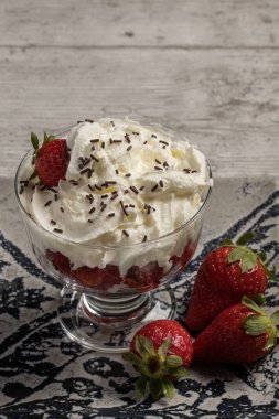 Süslü tabakta, ahşap zemin üzerinde kremalı ve çikolata parçalı lezzetli bir tatlı bardağı çileği. Dikey fotoğraf ve seçici odak