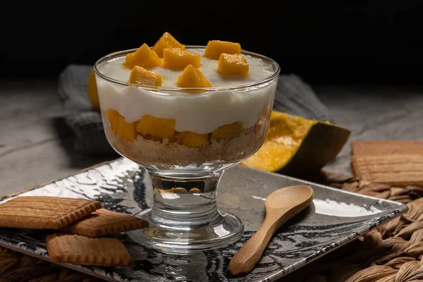 Mango ve bisküvi karışımlı lezzetli bir bardak soğuk yoğurt, siyah yemek tarzı. Tarif veya menü için yatay fotoğraf ve seçici odak
