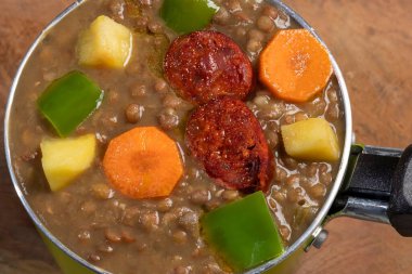 Stewed lentils with chorizo, apple or potato, green pepper and carrot in a green metal saucepan and on a wooden board. Concept of traditional and rustic cooking. selective focus