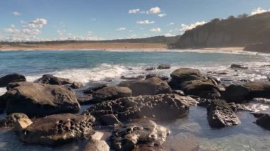 Okyanus dalgaları kayalara doğru yuvarlanıyor. Arka planda kumlu plaj var.