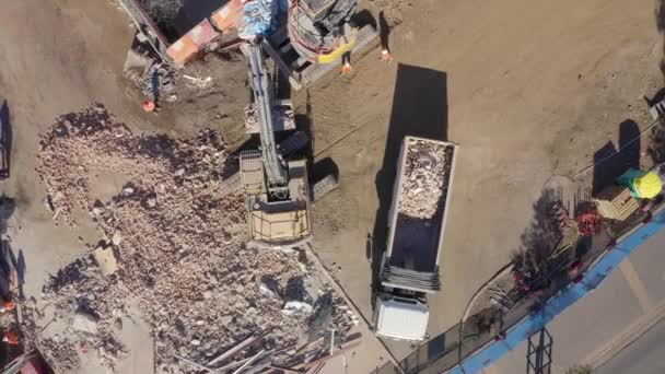 解体場所の空中ドローンビュー コンクリートおよび鋼鉄瓦礫をトラックに荷を積む掘削機 セーフティギアの労働者 — ストック動画
