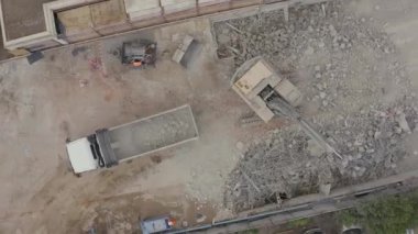 Yıkım alanının hava aracı görüntüsü. Kazıcı kamyona beton ve çelik moloz yüklüyor. Güvenlik donanımlı işçiler.