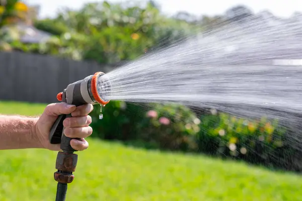 Garden hose with adjustable nozzle. Man\'s hand holding spray gun and watering plants, spraying water on grass in backyard.