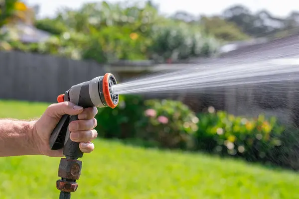 Garden hose with adjustable nozzle. Man's hand holding spray gun and watering plants, spraying water on grass in backyard.