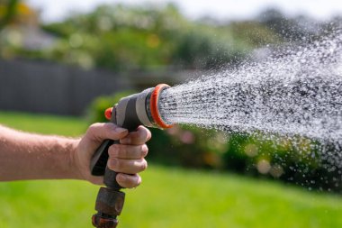 Ayarlanabilir hortumlu bahçe hortumu. Sprey tabanca tutan, çiçekleri sulayan arka bahçedeki çimlere su püskürten bir adam..