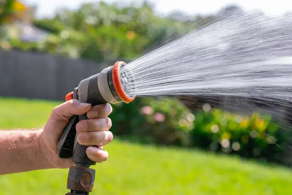 Garden hose with adjustable nozzle. Man\'s hand holding spray gun and watering plants, spraying water on grass in backyard.