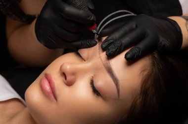 Beautician in black gloves making permanent makeup to a woman with a markup on a brows