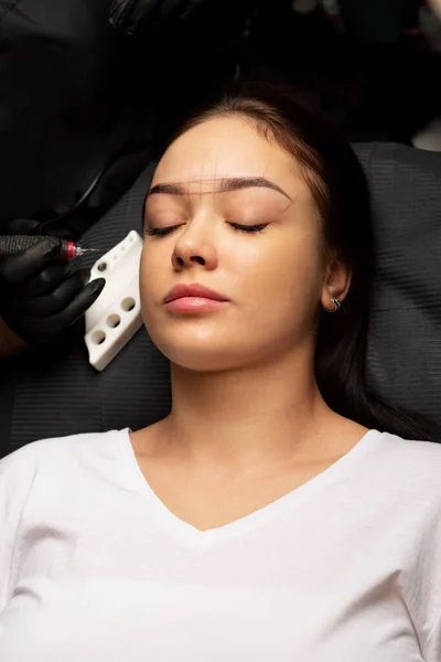 Beautician in gloves applying permanent brow makeup to a pretty brunette woman