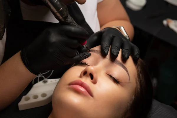 Cosmetician in gloves making permanent brow makeup to a woman at beauty salon. Closeup shot