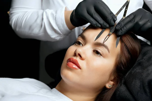 Cosmetician measuring the angle and forming the shape of the brows during the permanent tattoo procedure