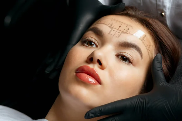 Brow master measuring the angle and forming the shape of the eyebrows during the permanent makeup procedure. Closeup shot