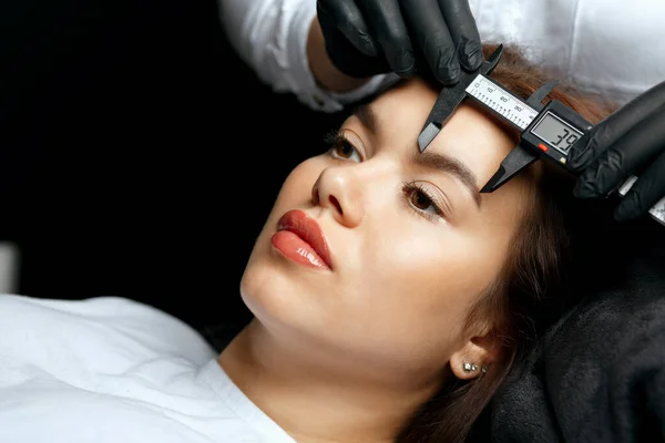 Beauty master measuring the angle and forming the shape of the eyebrows during the permanent makeup procedure