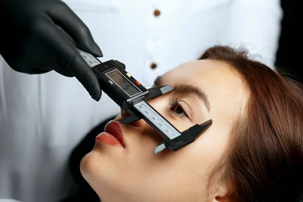 Brow specialist measuring the angle and forming the shape of the eyebrows during the permanent makeup procedure