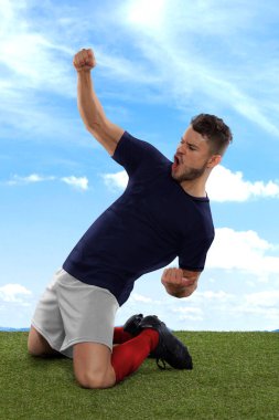 Mavi Fransa milli takımı forması olan profesyonel futbolcu sahadaki çimenler ve bulut arka planında meydan okuma ve mutluluk ifadesiyle gol attığı için heyecanla bağırıyor.