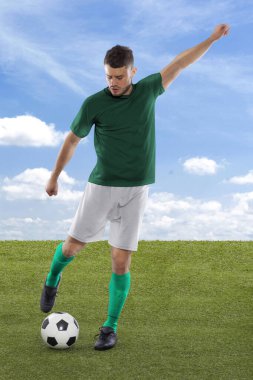 Yeşil Suudi Arabistan milli takımının formasına sahip profesyonel futbolcu, sahadaki çimenler ve bulut arka planında meydan okuma ve karar ifadesiyle gol atmak üzere.