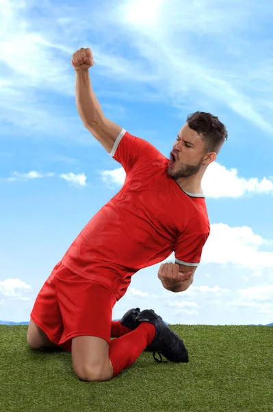 一名职业足球运动员 穿着红色的加拿大国家队球衣 兴奋地大喊着 因为他在场上的草地和云彩背景下 用一种挑战和快乐的表情射门 — 图库照片