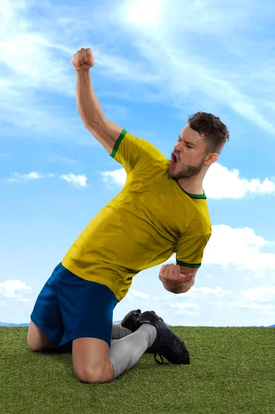 黄色のブラジル代表チームジャージを持つプロサッカー選手は フィールドグラスと雲の背景に挑戦と幸福の表現でゴールをマークするための興奮と叫んで — ストック写真