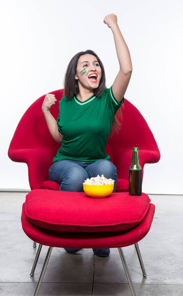 Красивая молодая женщина веер в зеленой футболке и флаге, нарисованный на лице сборной Саудовской Аравии празднует счастливое сидение в красном кресле с бутербродом и пивом на стуле.