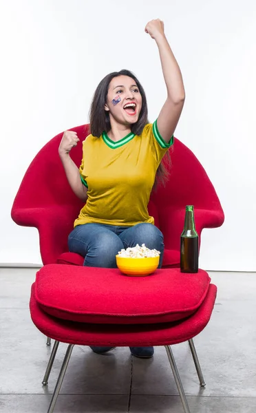 Beautiful young woman fan in a yellow t-shirt and flag painted on face of the Australia national team celebrating happy sitting in a red armchair with a sandwich and beer on a stool.