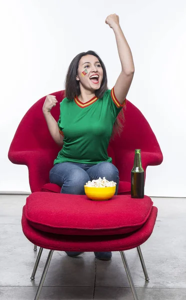 Beautiful young woman fan in a green t-shirt and flag painted on face of the Cameroon national team celebrating happy sitting in a red armchair with a sandwich and beer on a stool.