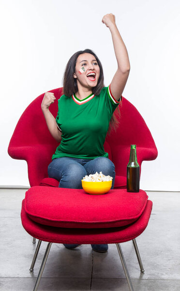 Красивая молодая женщина веер в зеленой футболке и флаге, нарисованный на лице сборной Мексики празднует счастливое сидение в красном кресле с бутербродом и пивом на стуле.