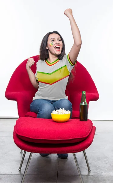 Beautiful young woman fan in a white t-shirt and flag painted on face of the Senegal national team celebrating happy sitting in a red armchair with a sandwich and beer on a stool.