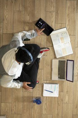 Young man with headphones sitting on the floor of his house studying and working at home with a computer, tablet, book, cell phone and glass of water.