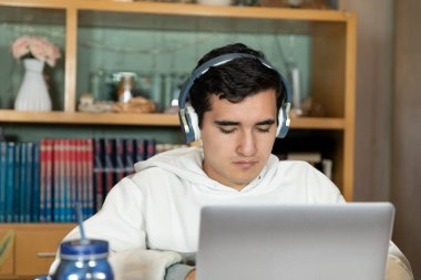 Young man with headphones sitting on the chair of his house studying and working at home with a computer, tablet, book, cell phone and glass of water. Home work.