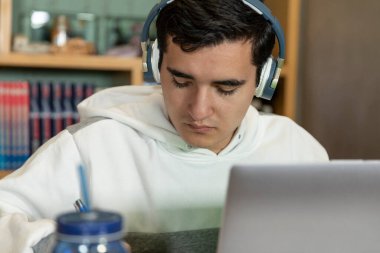 Young man with headphones sitting on the chair of his house studying and working at home with a computer, tablet, book, cell phone and glass of water. Home work.
