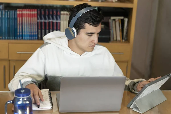 Young man with headphones sitting on the chair of his house studying and working at home with a computer, tablet, book, cell phone and glass of water. Home work.