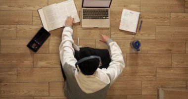 Young man with headphones sitting on the floor of his house studying and working at home with a computer, tablet, book, cell phone and glass of water.